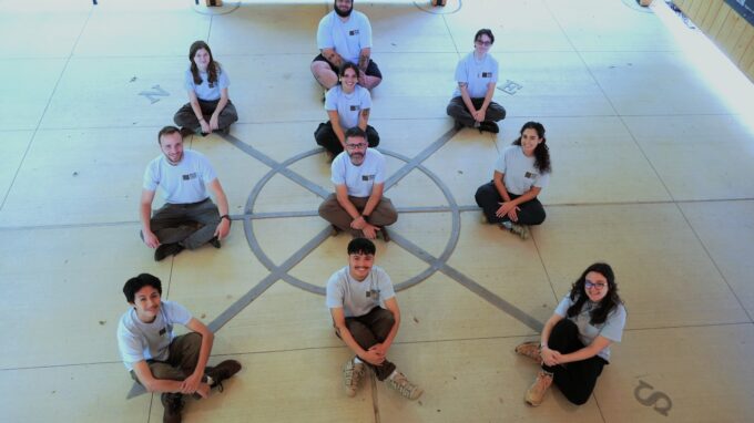 Nature Center education team sitting cross-legged on points of the compass placed on the ground of our Welcome Center. The team is all looking up at the camera and smiling.